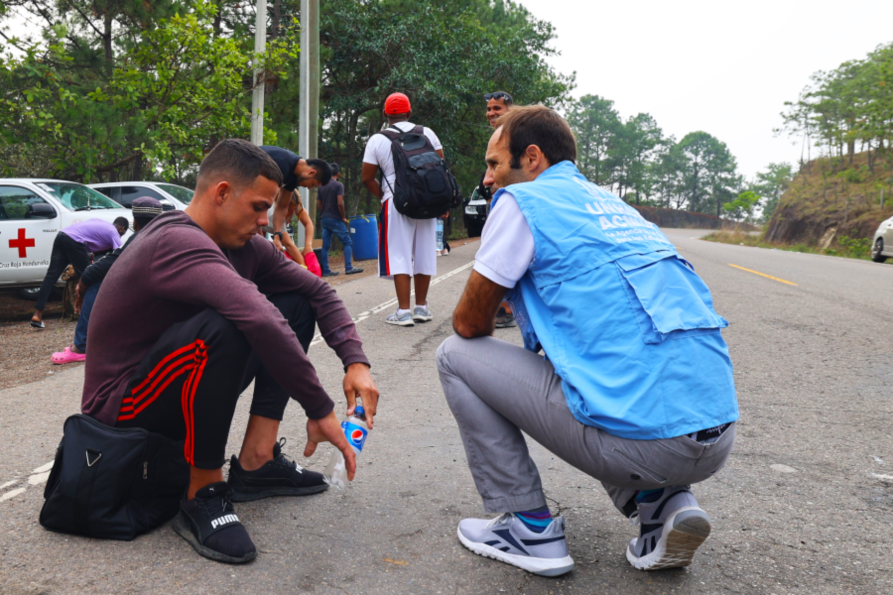 man in blue vest kneels down to speak to a young man sitting on the ground