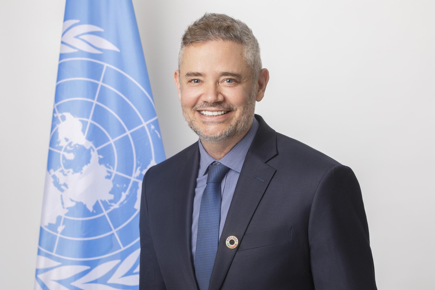 man in dark suit stands next to UN flag