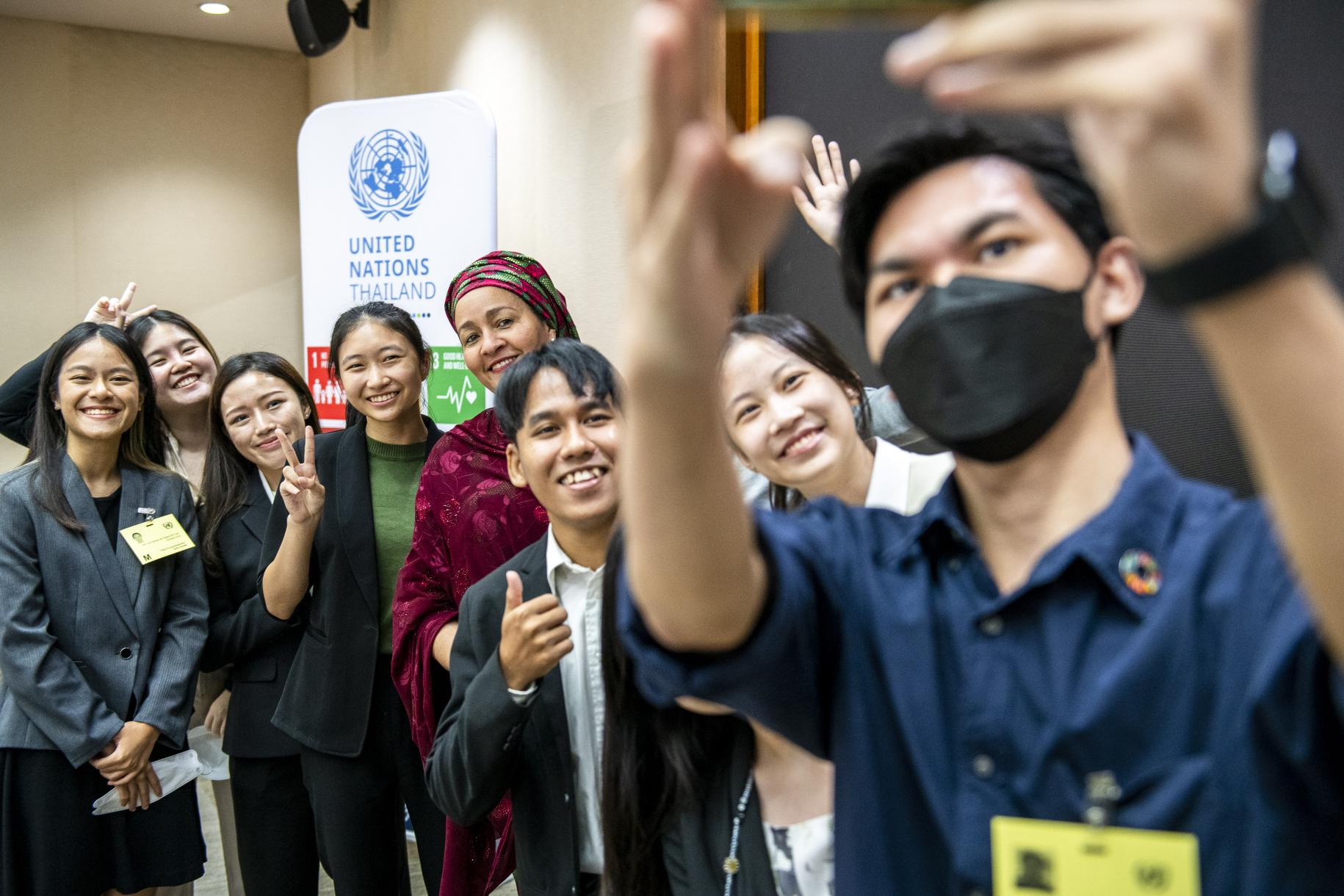 group of young people take selfie photo with woman in green and red headscarf in front of UN banner