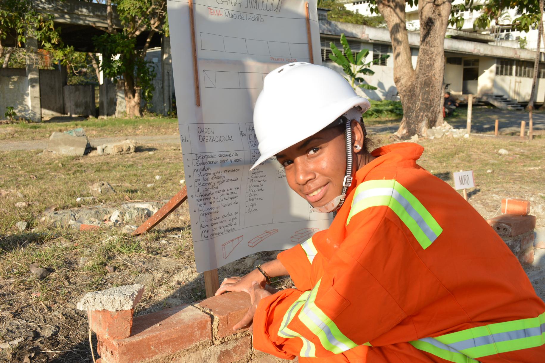 young person in hard hat and orange overalls lays bricks 