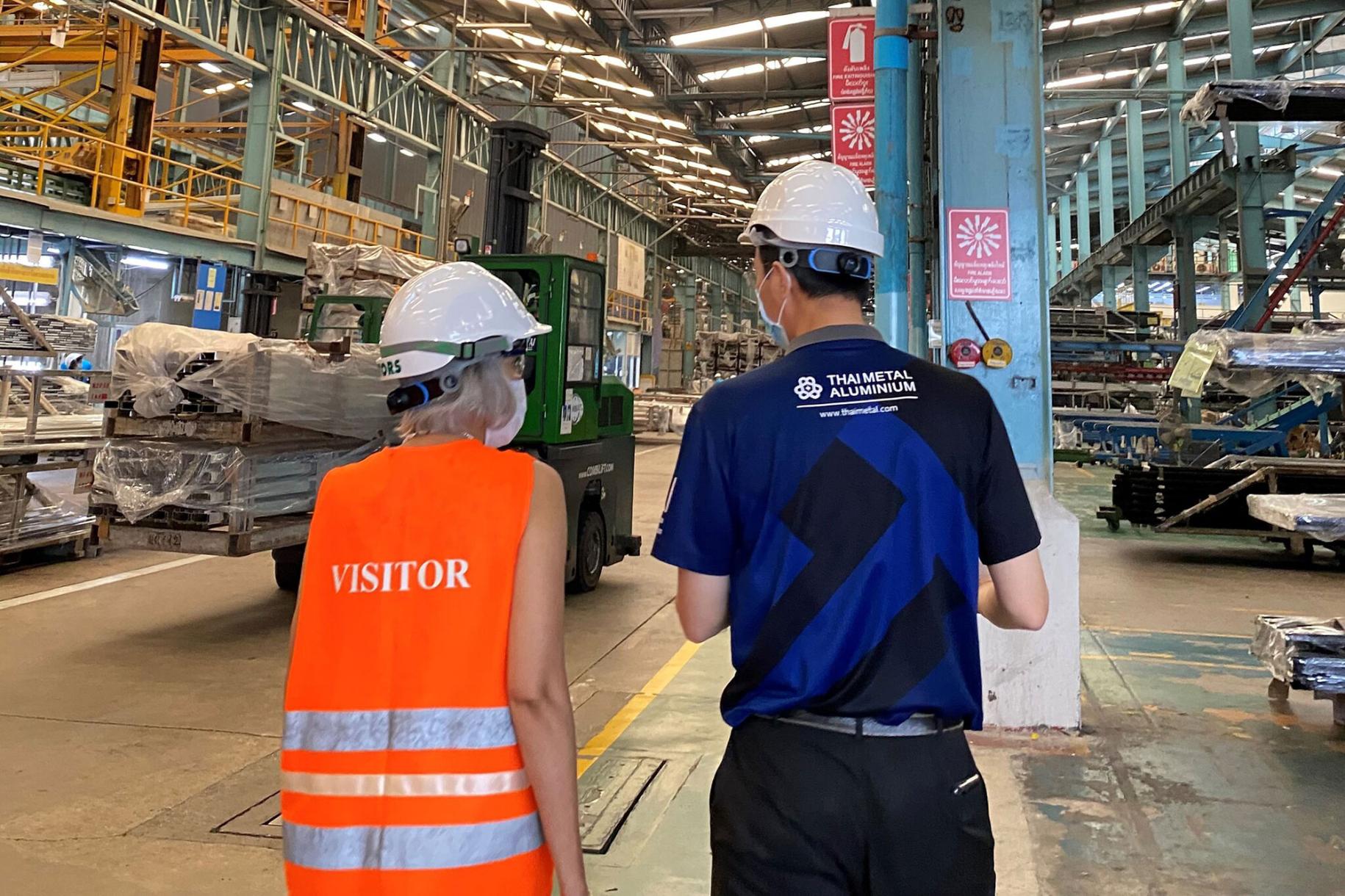 woman in orange vest and hard hat speaks to man in hard hat in large factory warehouse