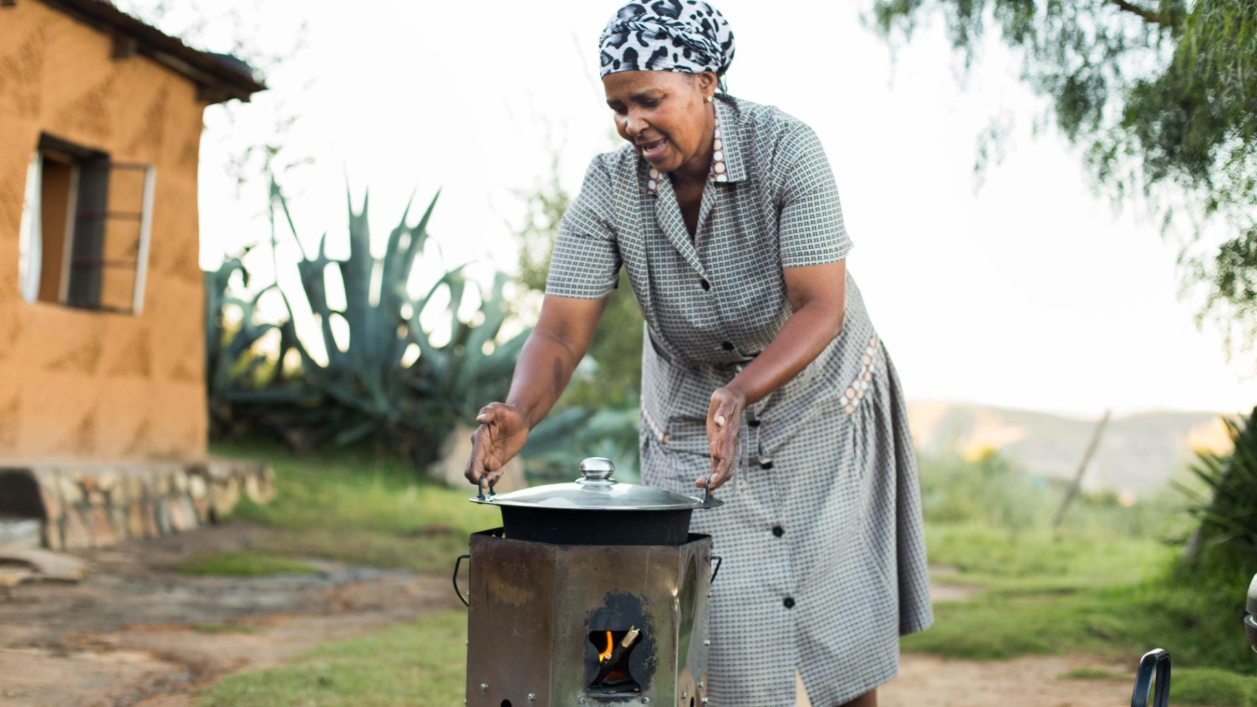 woman in dress and head scarf bends over outside stove to cook 