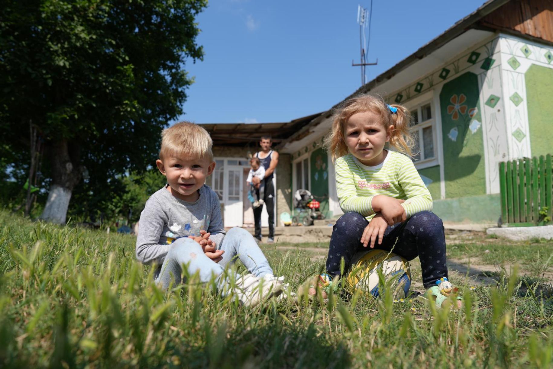 a young boy and girl sit on the grass outside their home, smiling at the camera