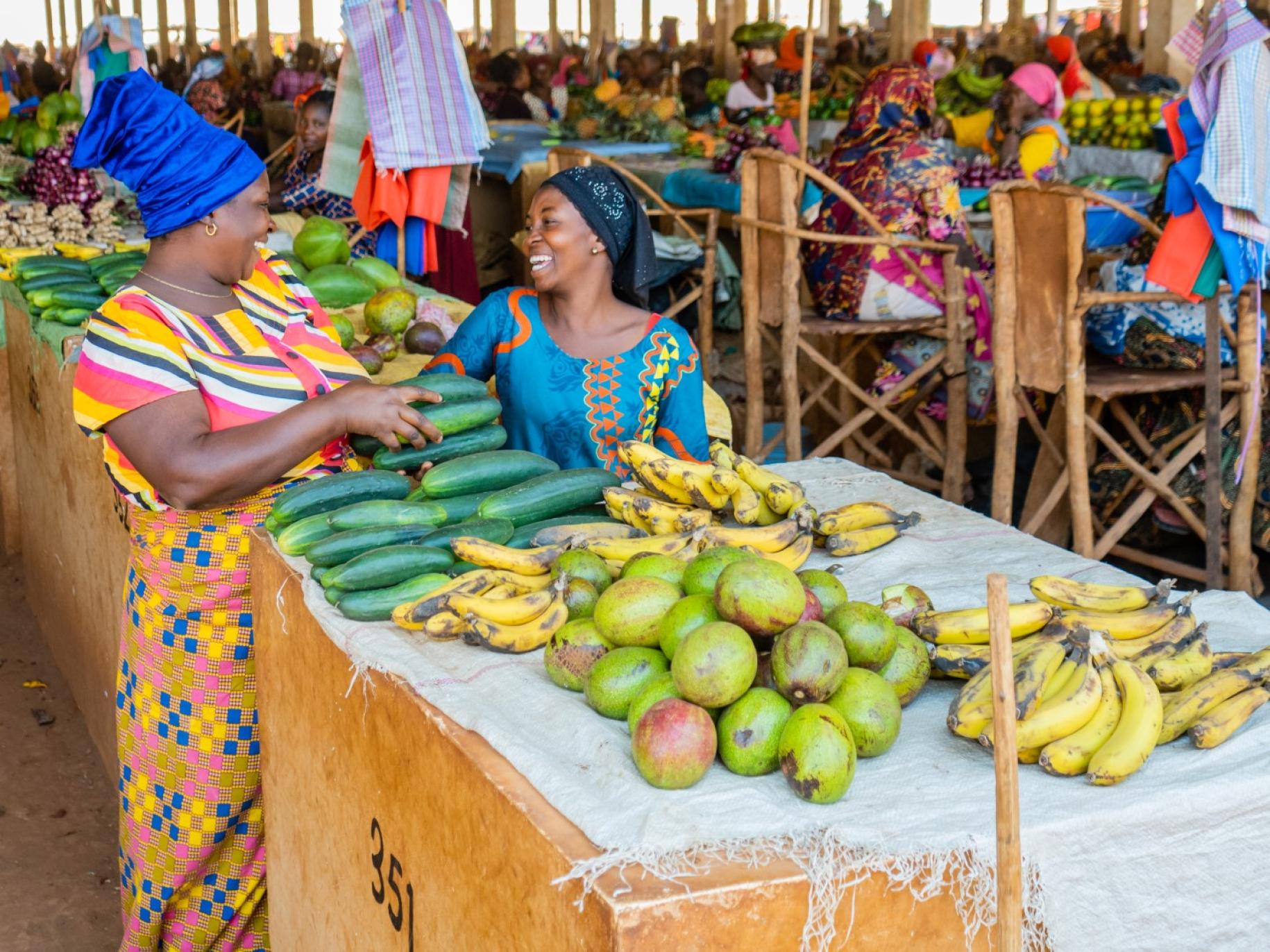 two women in bright clothes stand behind a mango and banana stand 