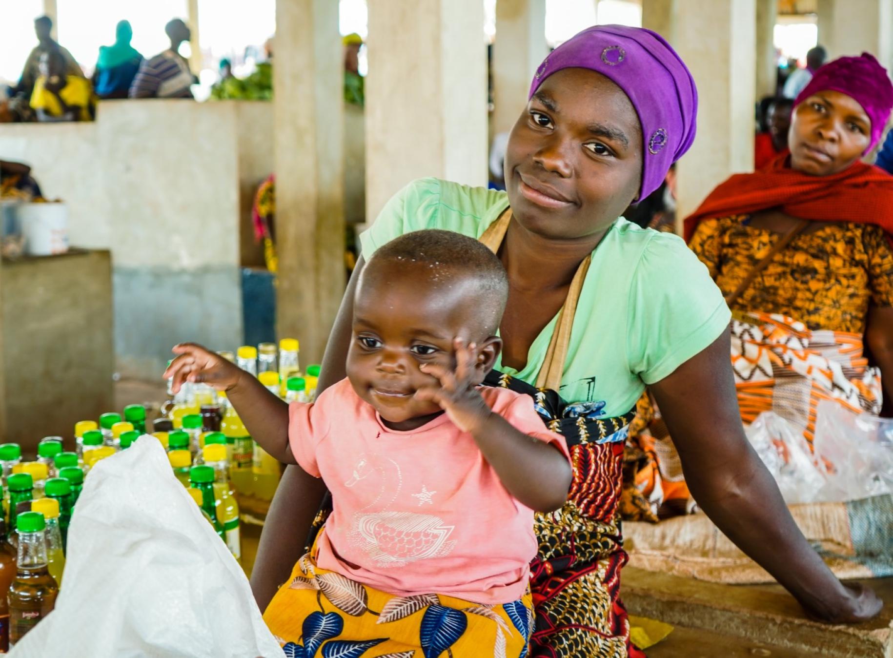 woman in purple head scarf sits in a market with child on her lap.