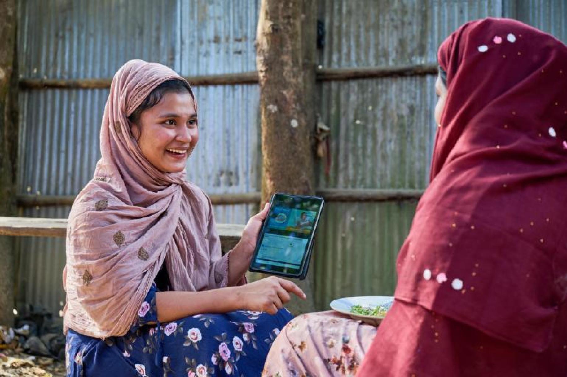 two young women in headscarves sit talking to one another holding a tablet