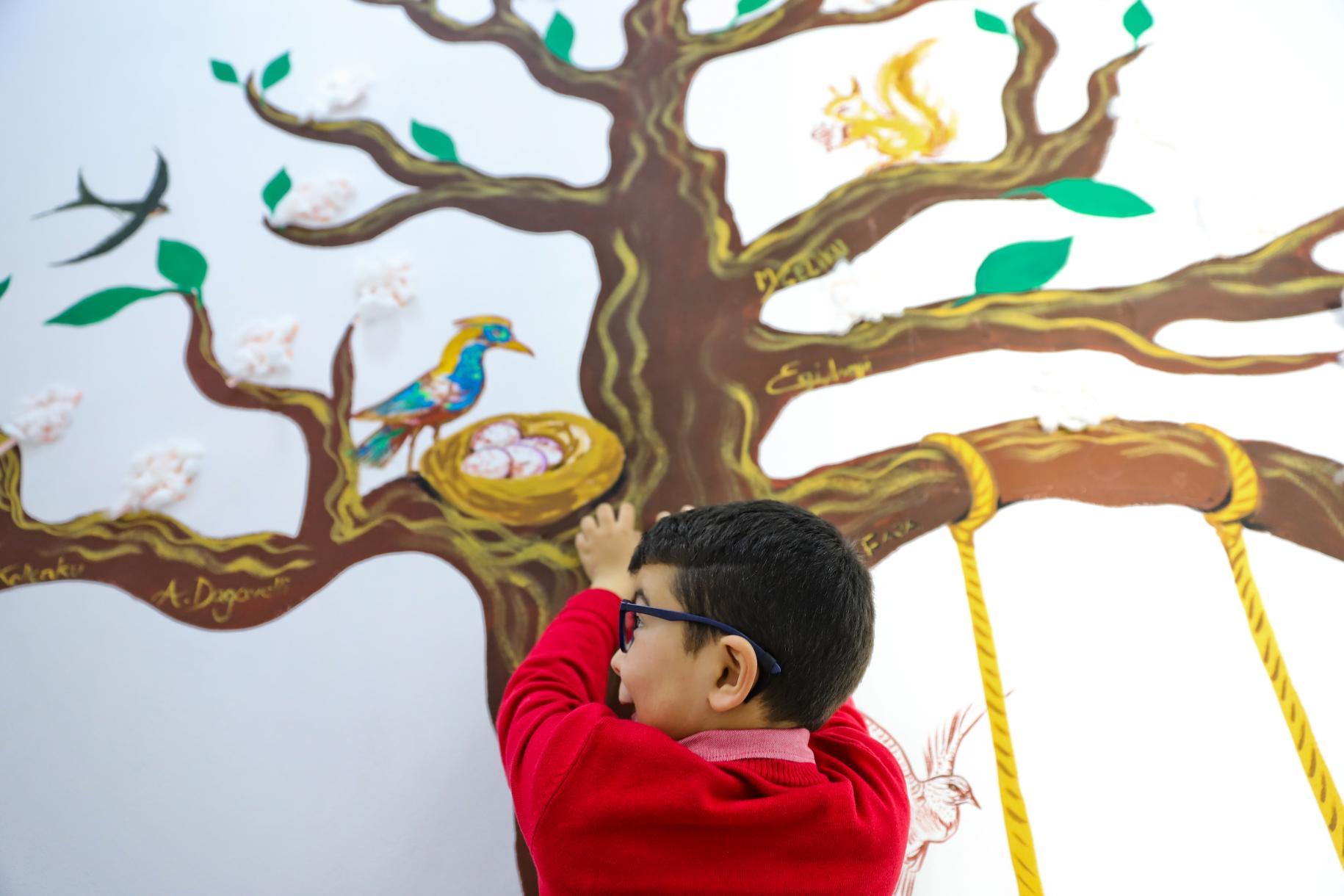 young boy in red top looks up to painting of a tree on the wall 