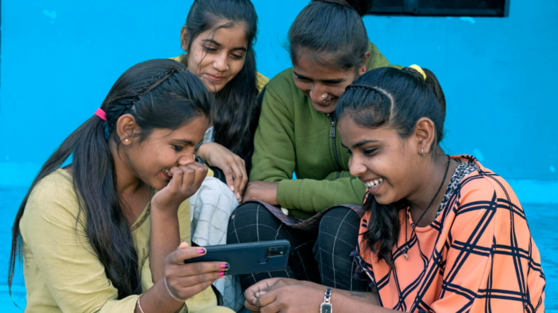 group of young women sit together against blue background looking at tablet