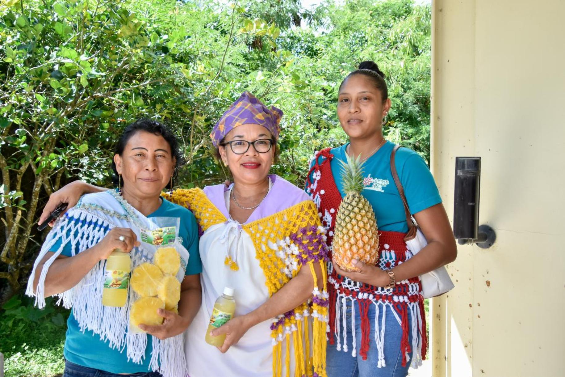 three women stand smiling towards the camera holding pineapples in their hands
