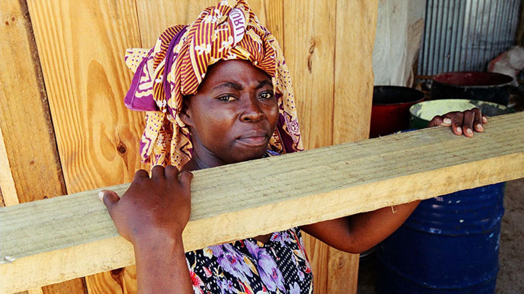 Women in colourful headscarf holds plank of wood on her shoulder and looks to the camera 