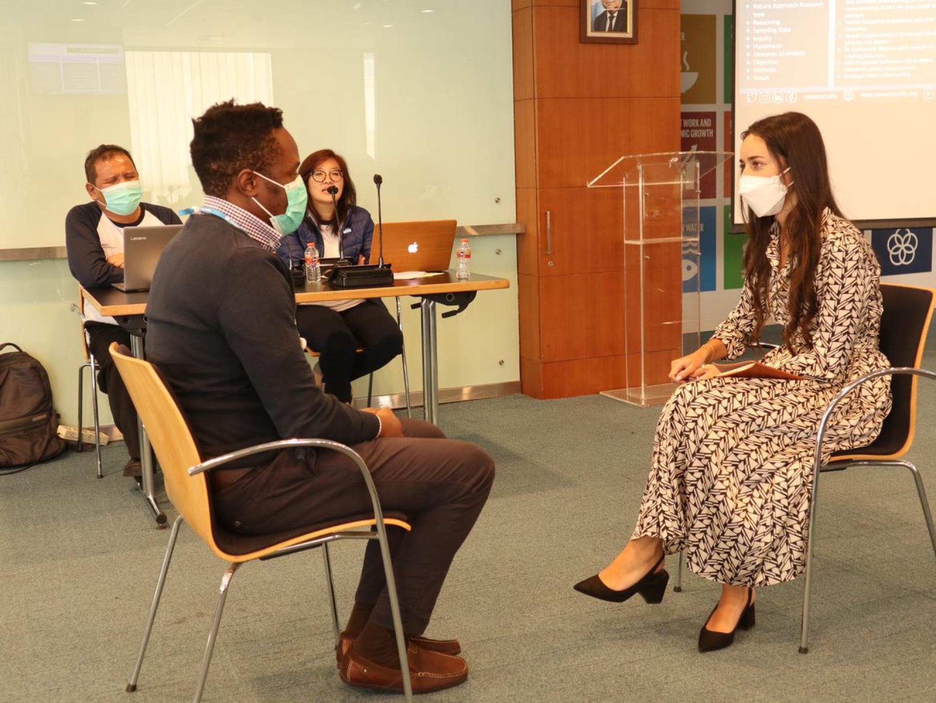 : Two people sit across from each another in a meeting room while two other people sit to their side at a separate desk
