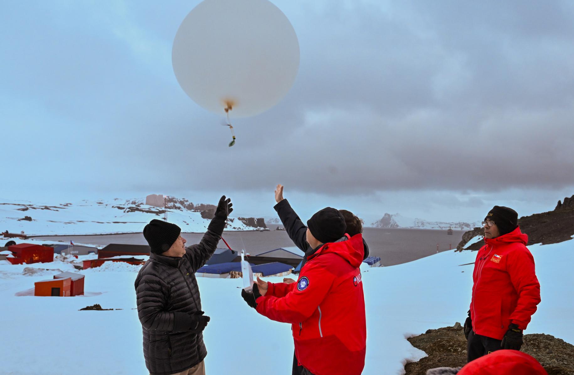 three men in waterproof clothing and hats stand on ice covered ground releasing a balloon into the air