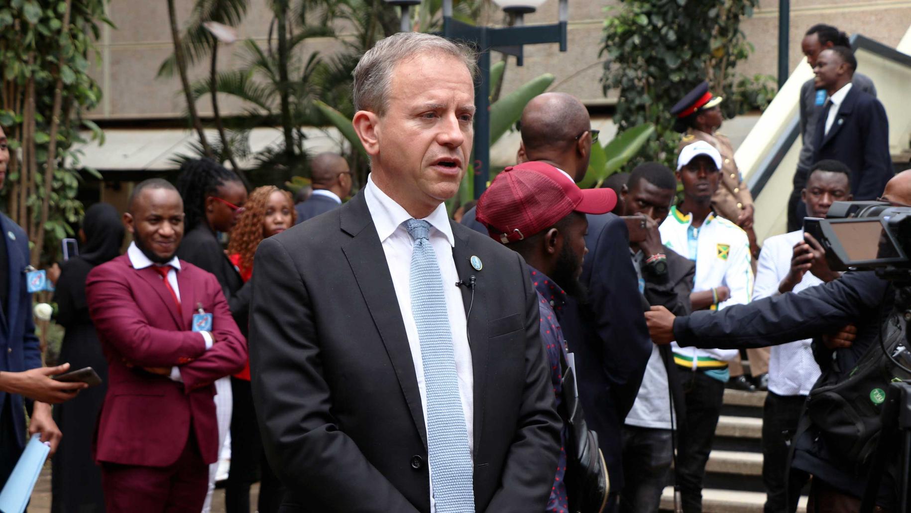 man in dark suit and blue tie stands side on to the camera with other people in the background