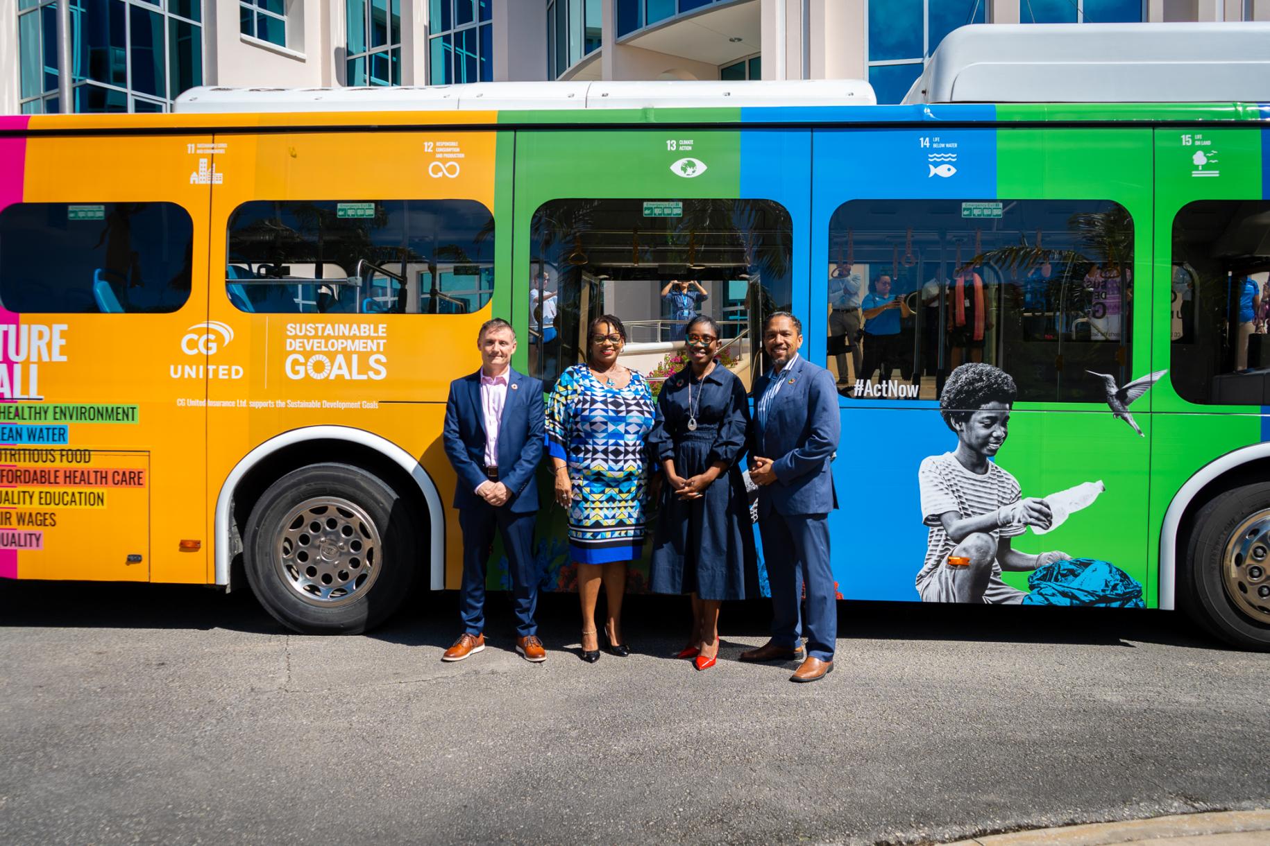 Four people, two men and two women in dark suits stand in front of a multicoloured bus