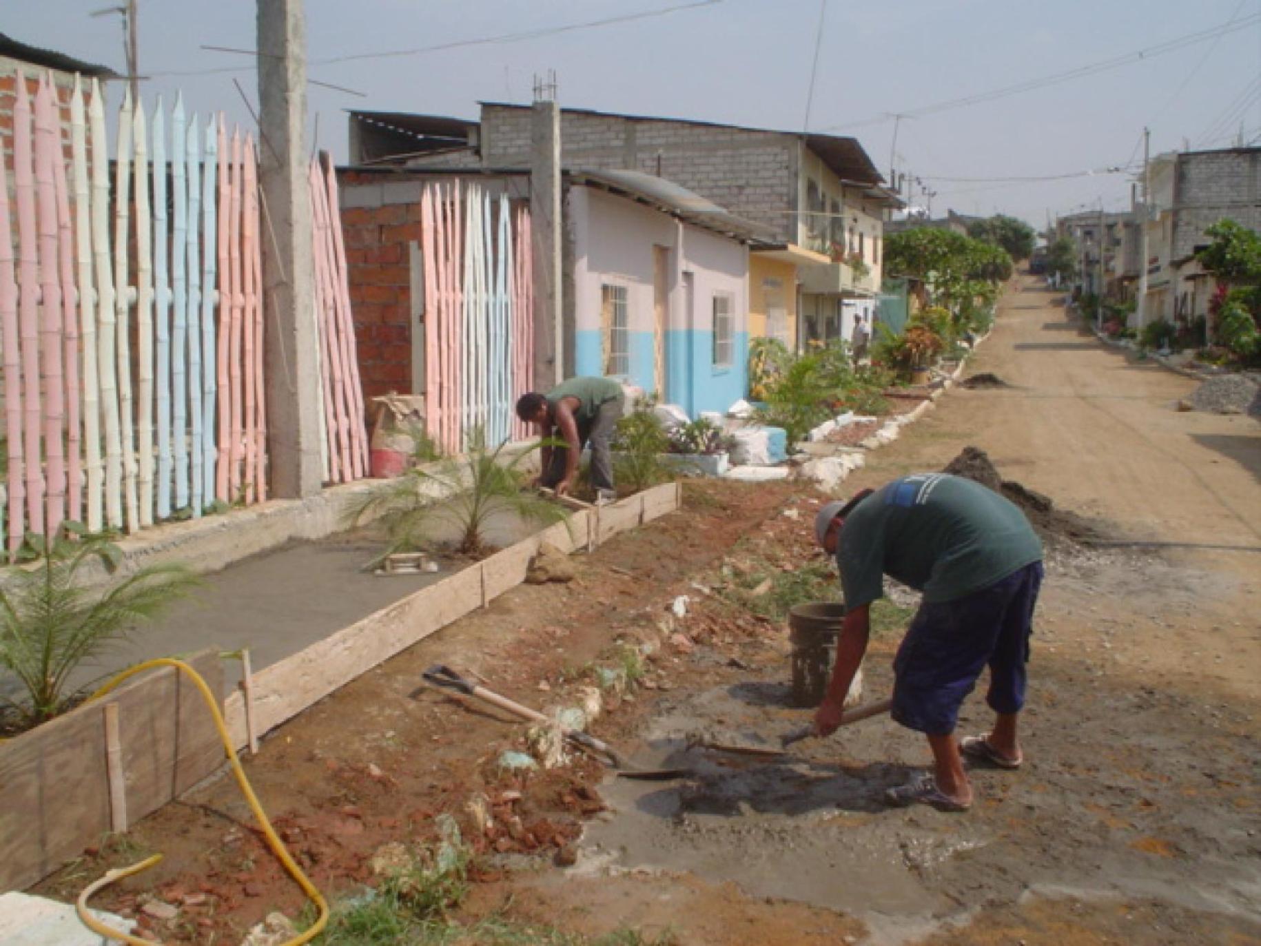 People plant plants outside colorful small buildings and a dirt road.