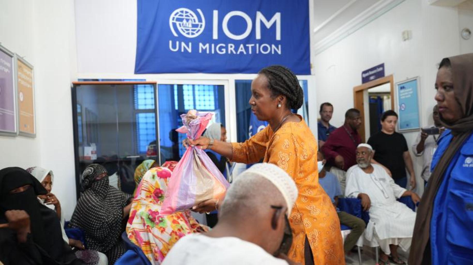 Men and women sit at a camp center in Sudan, with a big IOM UN Migration sign behind them. 