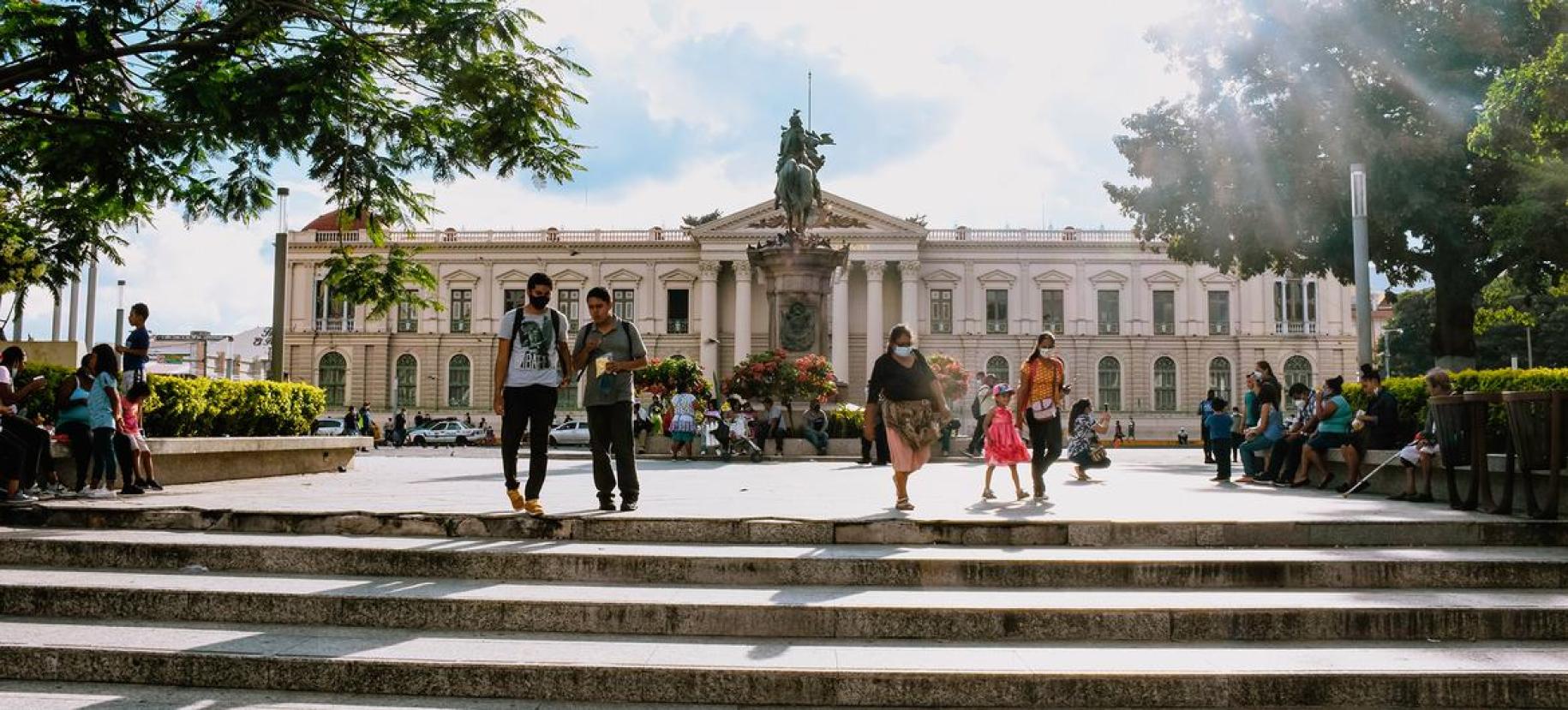 The image depicts a vibrant and sunny day at Gerardo Barrios Square in El Salvador. People are seen walking, standing, and socializing in the foreground. Steps lead up to the main area of the square where more people are gathered. 