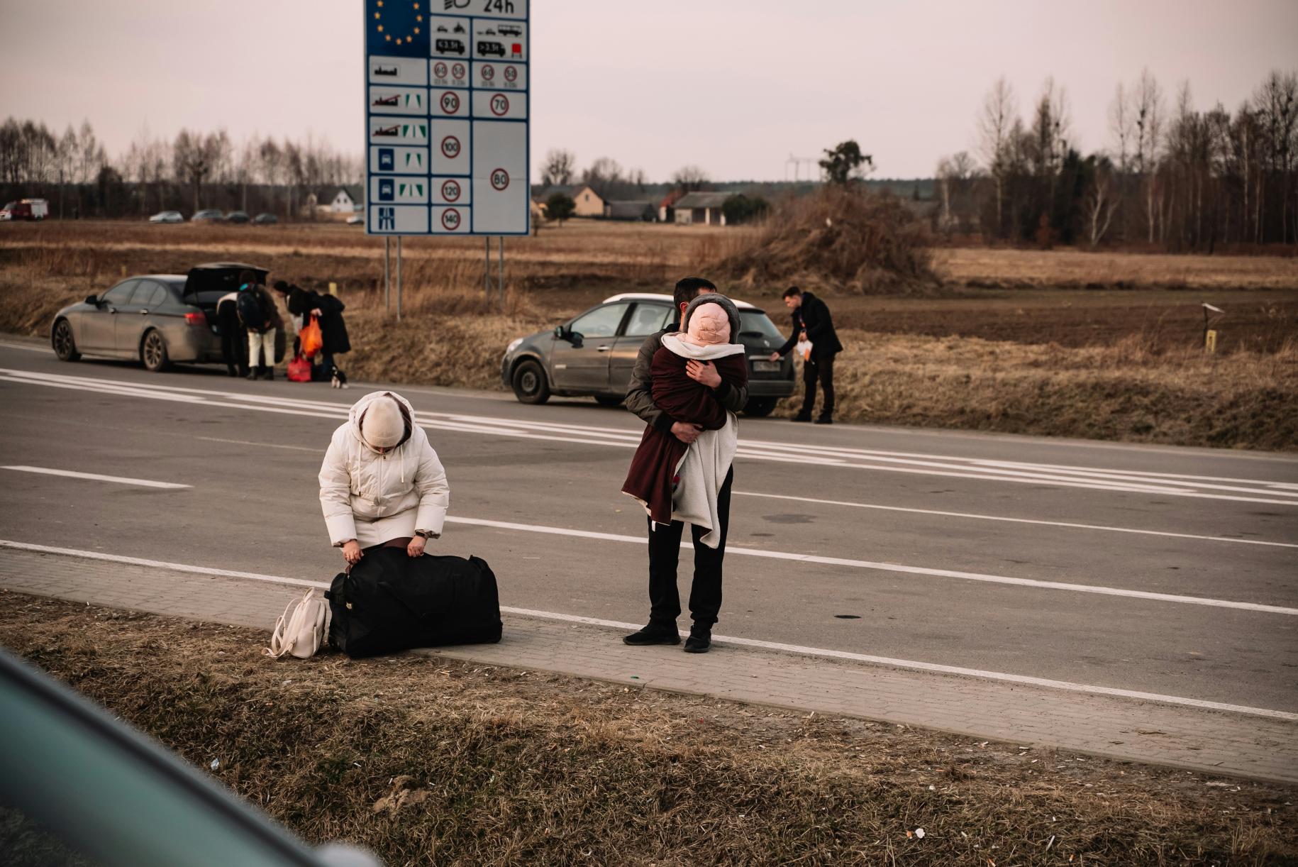 A woman sits on the side of the road with her luggage, while a man carries a child and cars pass by