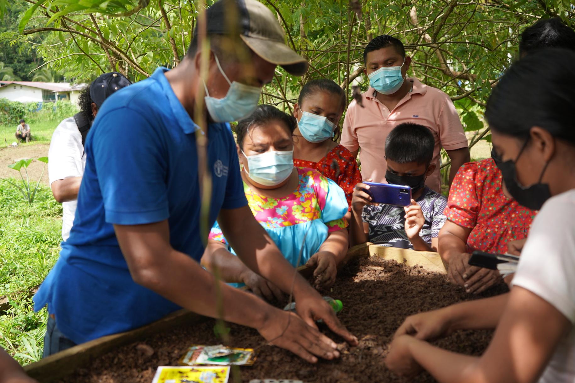 A man wearing an FAO shirt is planting seedlings, while a group of women look on