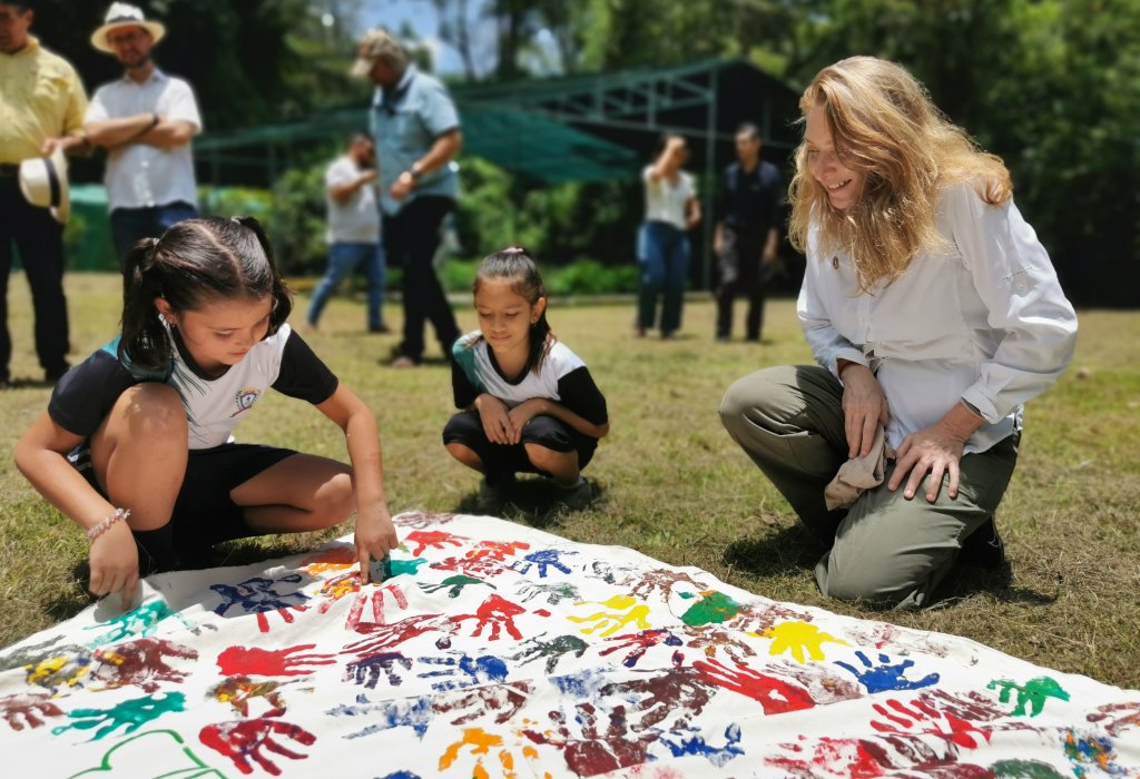 Two children and UNRC are kneeling on the grass beside a large white cloth spread out on the ground.