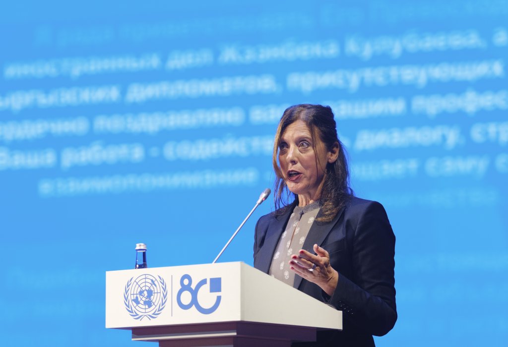 A woman stands at a podium with a UN emblem, delivering remarks at a formal event against a blue backdrop.