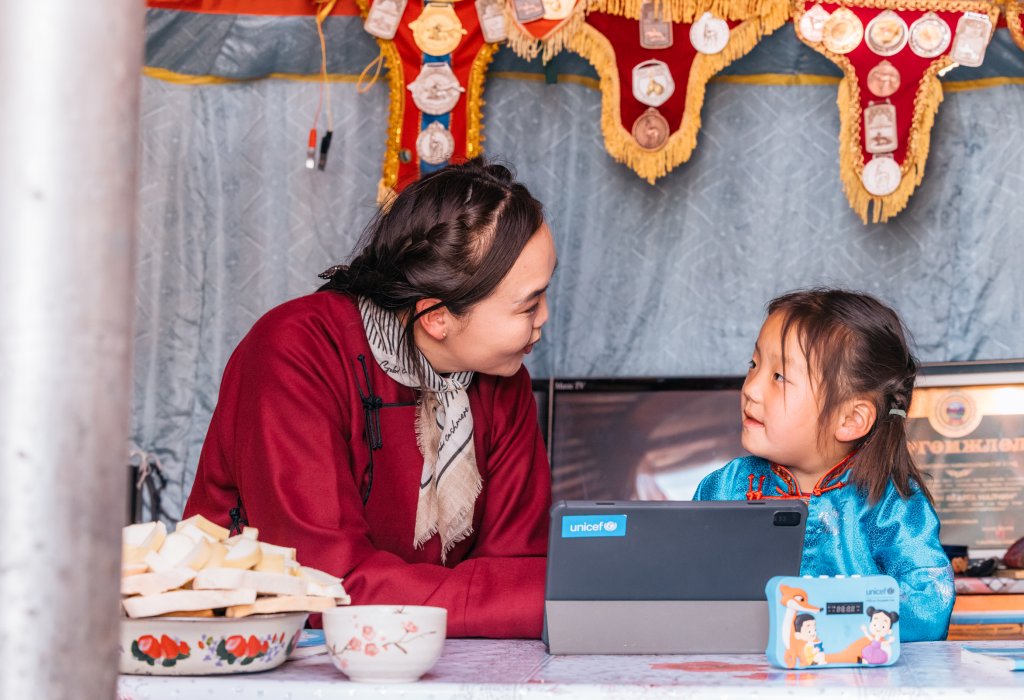 A teacher and a young girl share a warm moment over a tablet inside a traditional ger.