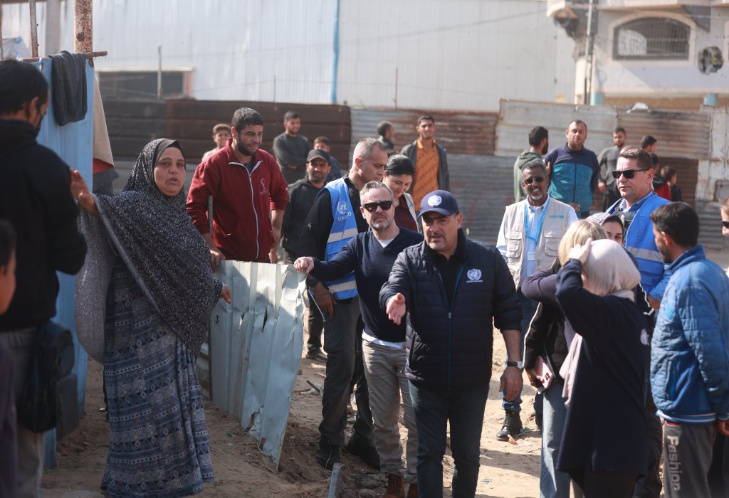 A group of people, including individuals in UN-marked vests, engage in a discussion with community members in a settlement area with makeshift structures.