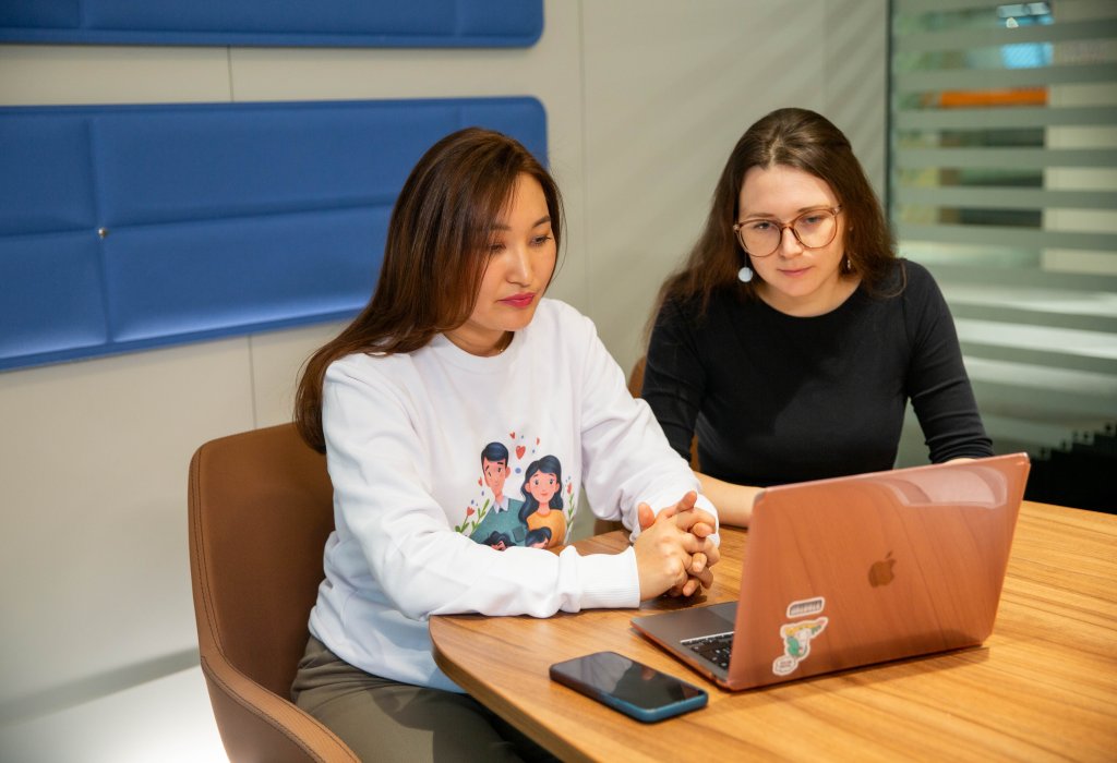 Two people sit at a table in a meeting room, looking at a laptop together with a phone nearby.