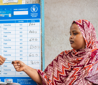 Two women interacting in a fish market. Both are wearing hijabs and traditional African tunics.