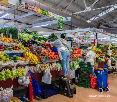 Busy indoor market stall displaying colorful fruits and vegetables, with shoppers browsing and a vendor serving produce.