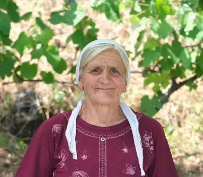 A woman in Albania wearing a mauve dress and a white headscarf in front of a table with food.